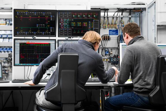 Fotografiert sind zwei männliche Personen. Sie sitzen mit dem Rücken zur Kamera an einem Schreibtisch. Vor ihnen befinden sich Monitore mit technischen Daten. Im Hintergrund sind Schaltkreise zu sehen.