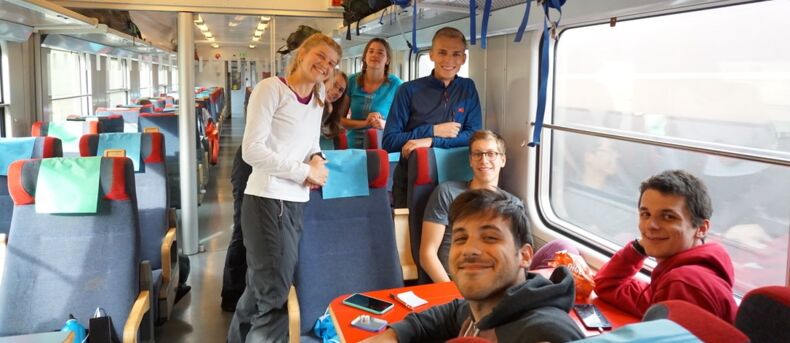 The picture shows seven people on a train. Three people are sitting at a table. Four people are standing. They are all smiling at the camera.