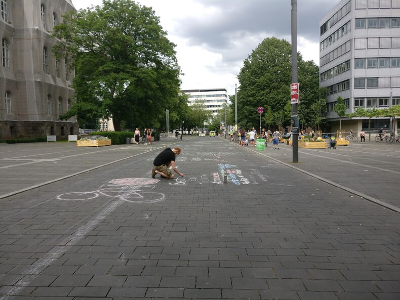 Das Bild zeigt eine Person, die auf dem Templergraben kniet und mit Kreide die Straße bemalt. Im Hintergrund sind Sitzgelegenheiten aus Holz auf dem Platanenplatz zu sehen.