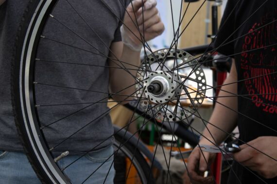 The picture shows the spokes of a bicycle in the foreground. In the background are two people, one holding a tool.