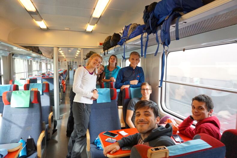 The picture shows seven people on a train. Three people are sitting at a table. Four people are standing. They are all smiling at the camera.