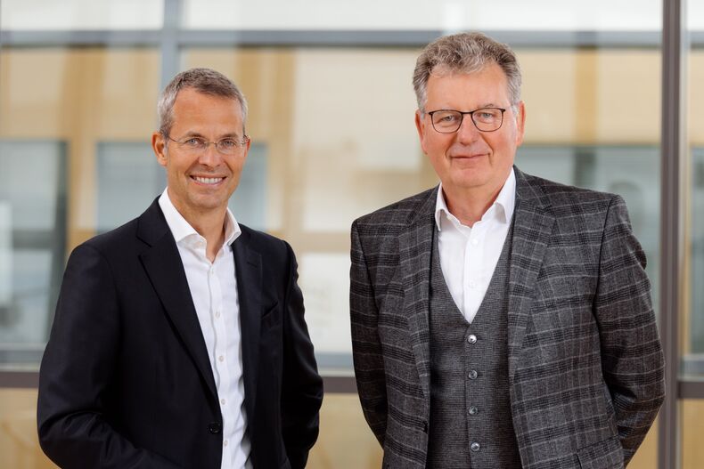 Die Prorektoren Professor Malte Brettel, Prorektor für Wirtschaft und Industrie und Professor Matthias Wessling, Prorektor für Forschung und Struktur, stehen vor einer Glaswand.  Professor Brettel trägt einen schwarzen Anzug mit weißem Hemd, Professor Wessling ein grau kariertes Sakko, eine graue Weste und Hose, sowie ein weißes Hemd.  Sie lächeln in die Kamera.