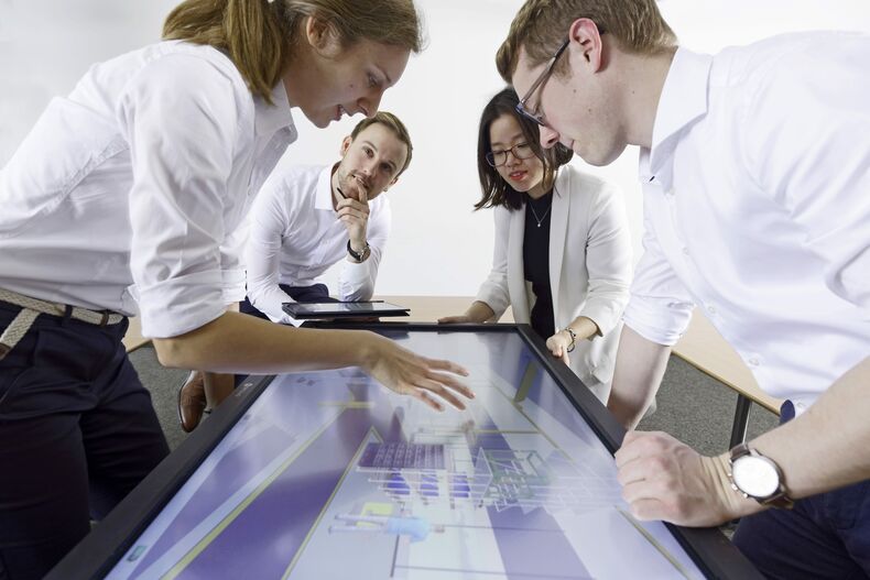 There are a total of four people in the photo, two women and two men. They are bending over a mo-nitor with a technical model.