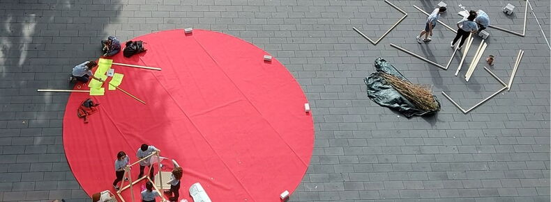 Das Bild zeigt eine Aufnahme aus der Vogelperspektive. Auf einem großen, runden roten Teppich bauen Teilnehmende der Summer School 2022 Kunstinstallationen auf und werden dabei gefilmt.