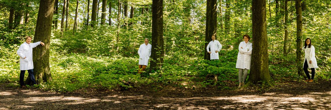 Zwei weibliche und drei männliche Personen in weißen Laborkitteln stehen in unterschiedlichen Posen an Bäume gelehnt verteilt im Wald. Sie lächeln in die Kamera.